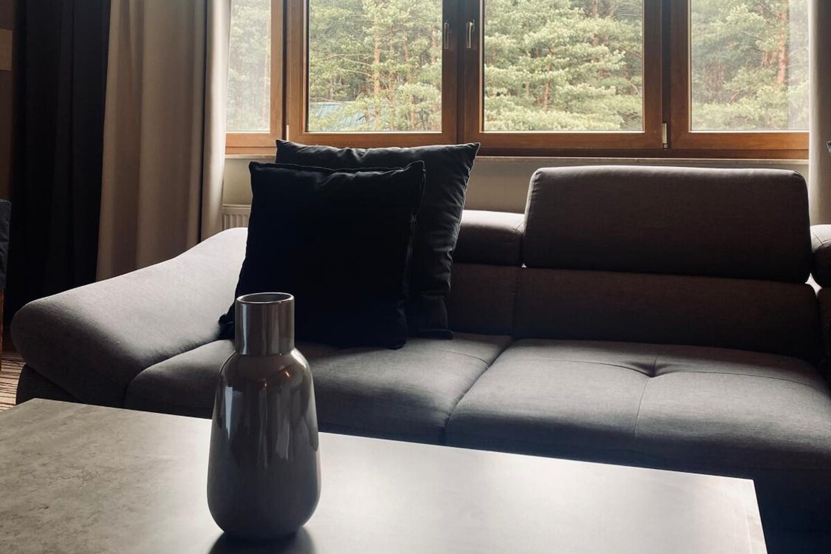 Een moderne woonkamer met een bank, een bijzettafeltje en een vaas. Grote ramen bieden uitzicht op een bebost gebied.
