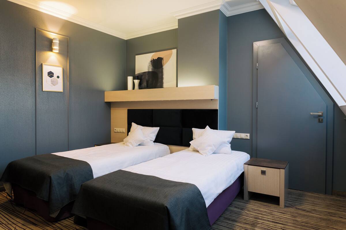 Une chambre d’hôtel avec deux lits, une petite table de chevet, une lampe fixée au mur et un tableau au-dessus du lit.