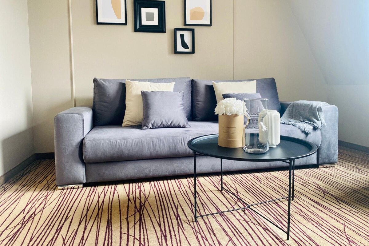 Een moderne woonkamer met een grijze bank, een ronde salontafel en een kamerbreed tapijt met een patroon.