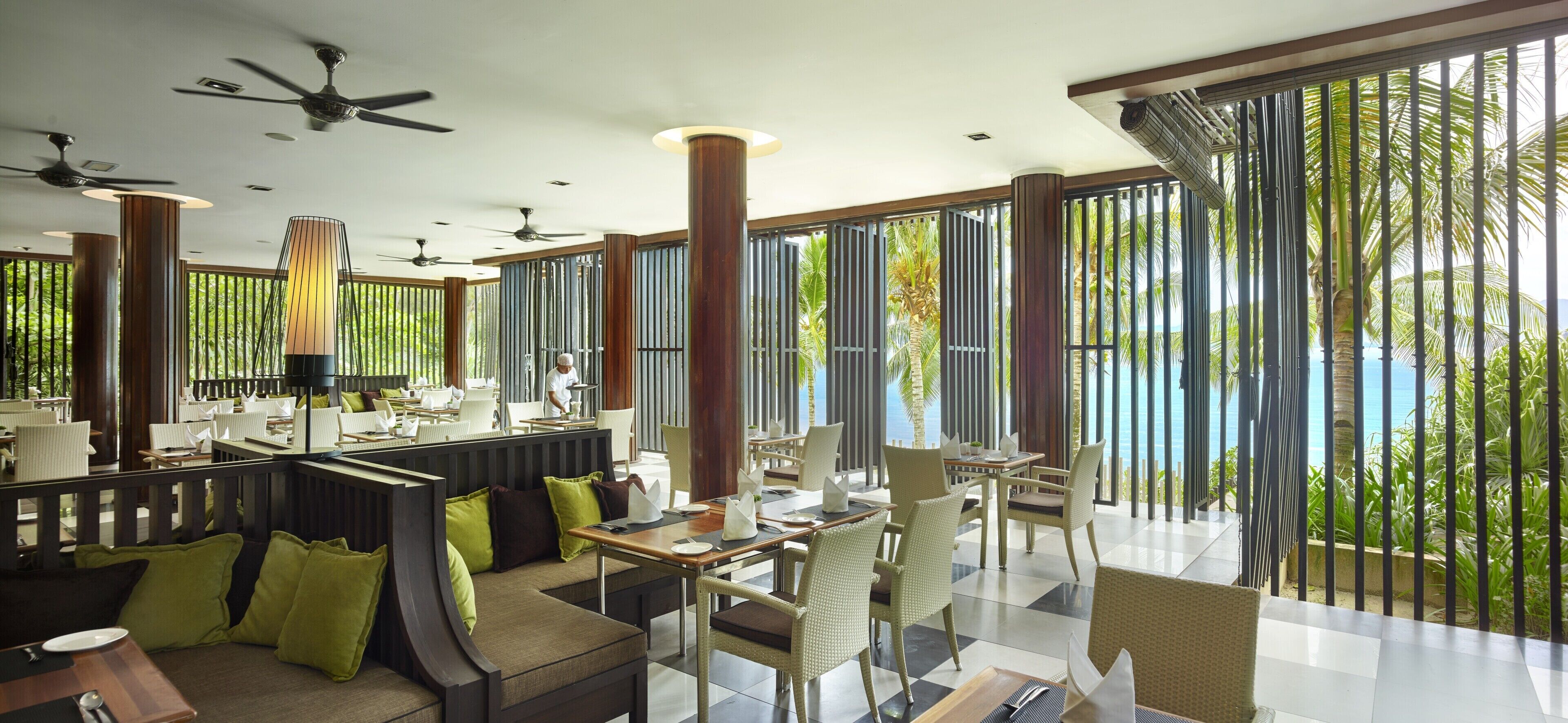 A modern restaurant with high ceilings, ceiling fans, and large windows offering a view of the ocean.