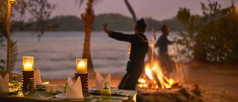A table set for a meal with candles, a fire in the background, and a beach view.