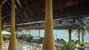 An outdoor lounge area with wooden pillars, tables, and chairs, overlooking a pool and the sea.