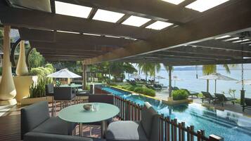 An outdoor pool area with lounge chairs and tables, a view of the sea, and a wooden deck.