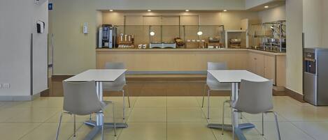 Una cafetería moderna con dos mesas y sillas, una estación de autoservicio para bebidas y un refrigerador.