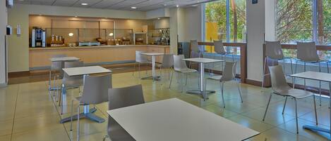 A spacious cafeteria with white tables and chairs, a counter with a coffee machine, and large windows offering a view of trees.