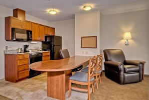 A compact kitchen with wooden cabinets, a microwave, and a refrigerator. There is a dining table with chairs and a single leather armchair. - Candlewood Suites Burlington South by IHG (Burlington)