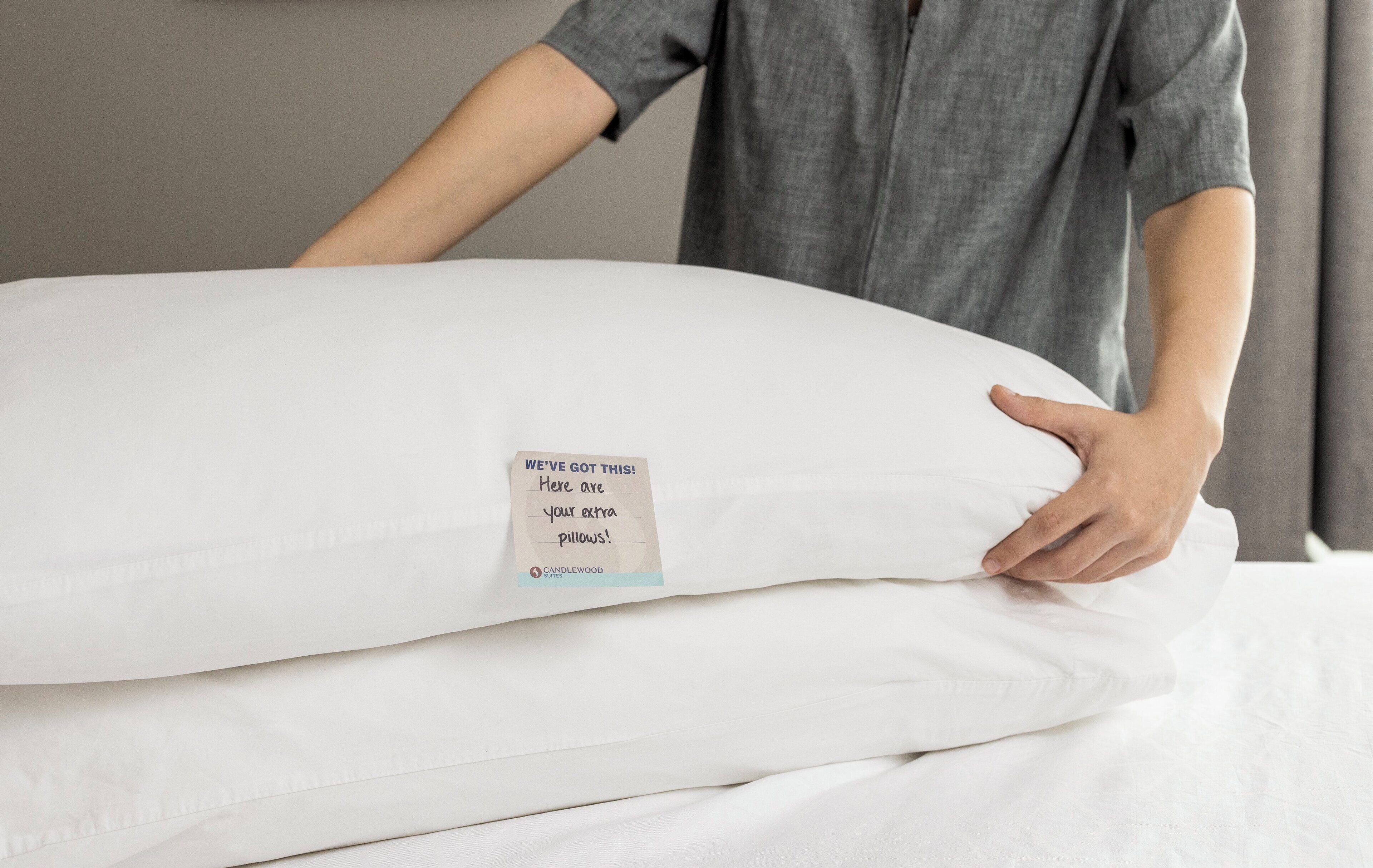 Una persona sostiene un almohadón blanco grande con una etiqueta que dice "¡NOSOTROS LO TENEMOS! Aquí tiene sus almohadas adicionales" y el logotipo de Candlewood Suites.