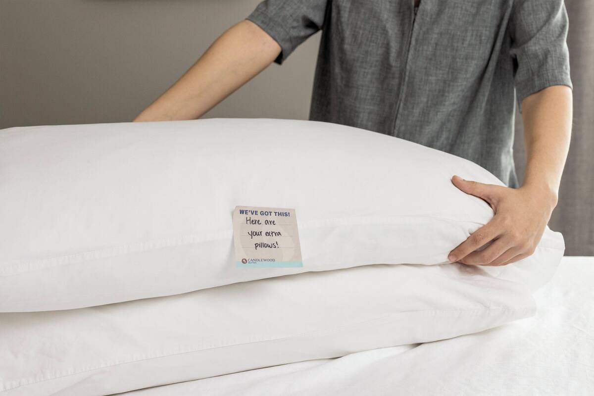 Une personne tient un grand oreiller blanc sur lequel est attachée une étiquette indiquant : « ON S’EN OCCUPE ! Voici vos oreillers supplémentaires ! » et sur lequel figure également le logo de Candlewood Suites.