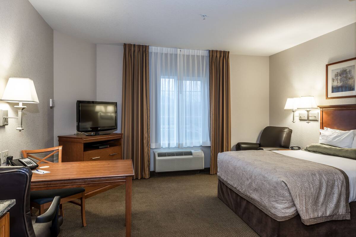 Une chambre d’hôtel comprenant un lit, un bureau avec une chaise, une télévision, une chaise, une lampe et une fenêtre avec des rideaux.