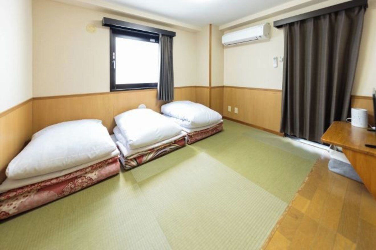 A hotel room with two beds, a small desk, a window with curtains, and an air conditioning unit.