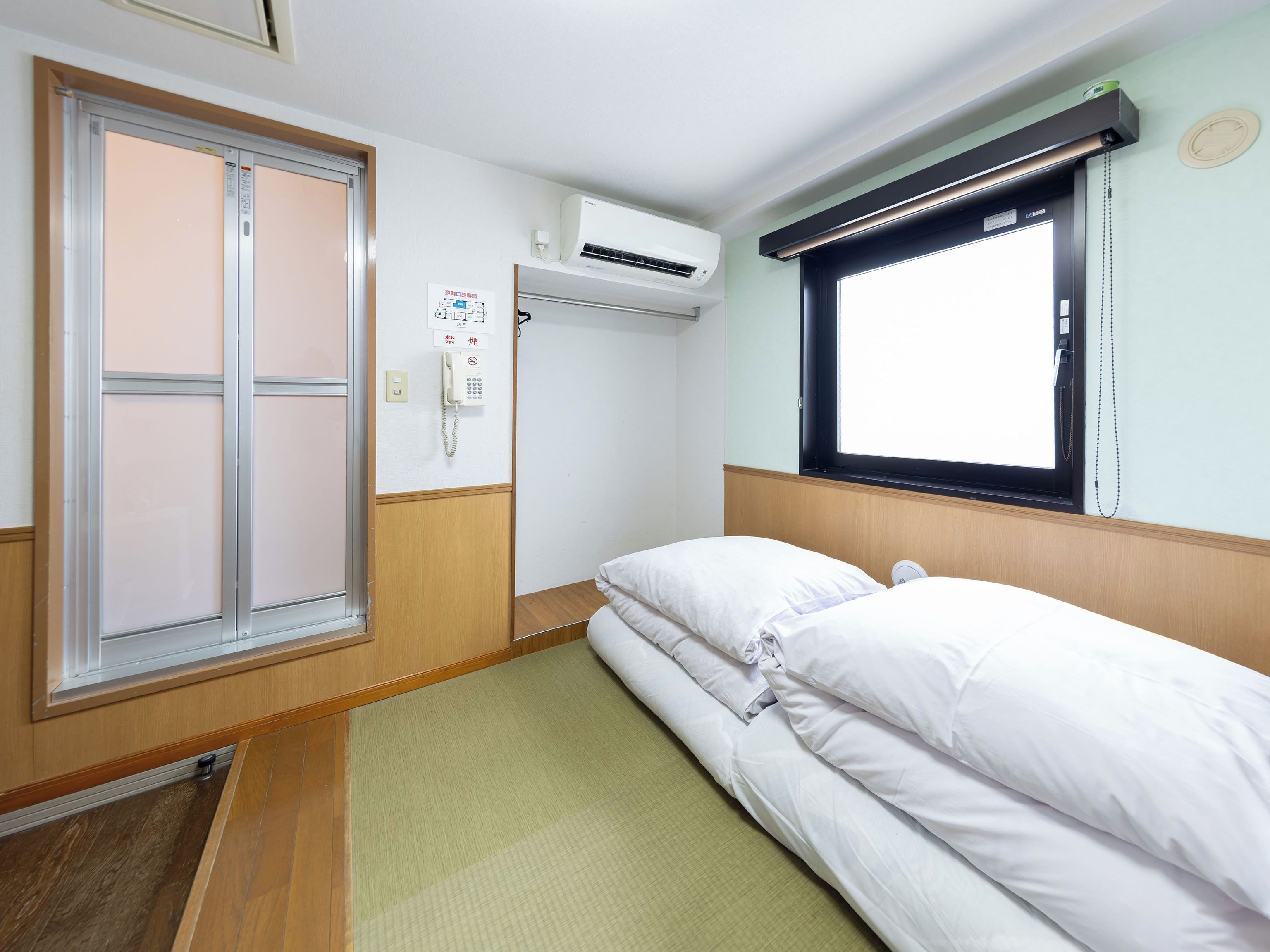 Habitación de hotel con cama, ventana con persianas, unidad de aire acondicionado y teléfono.