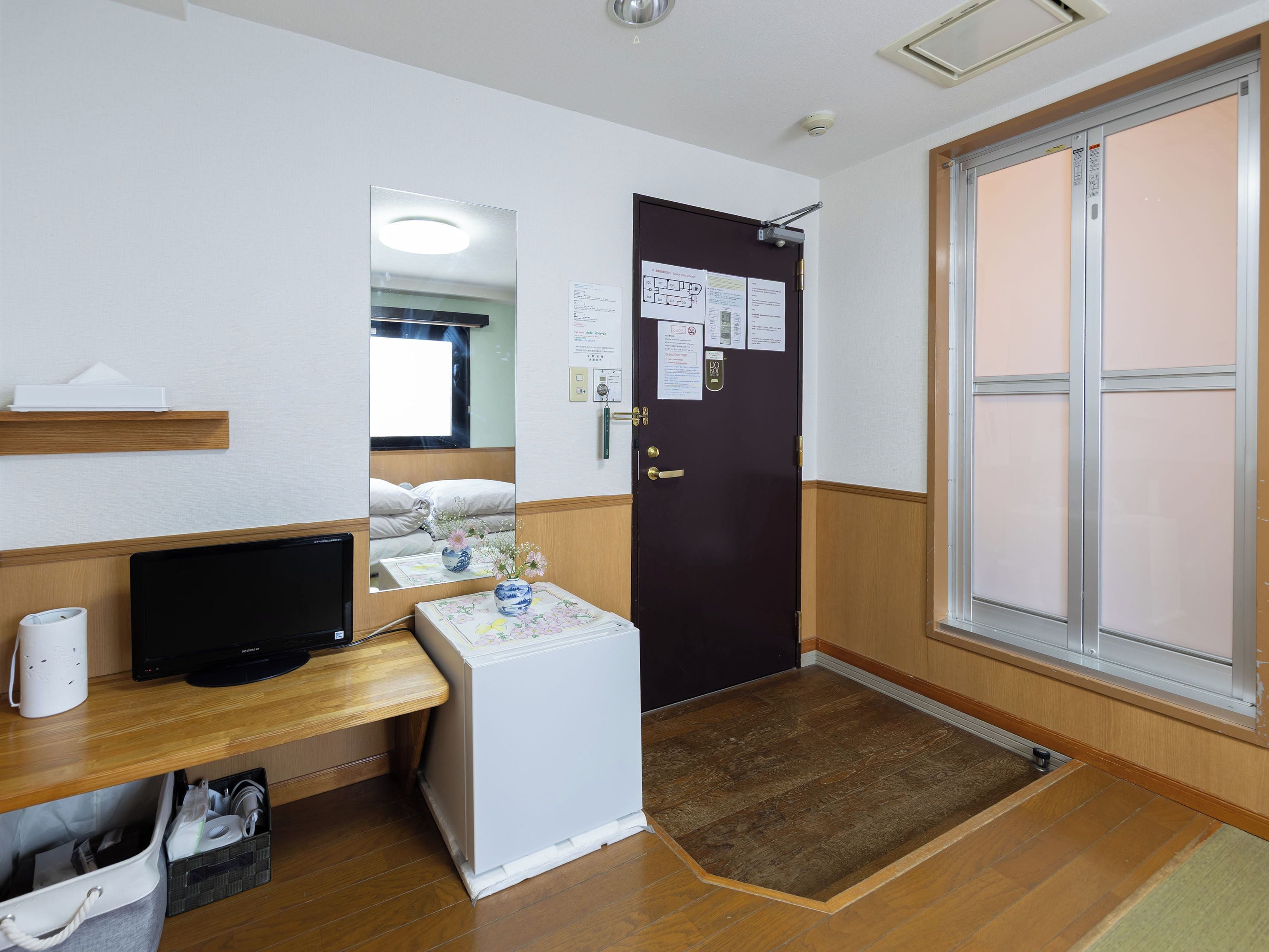 A hotel room with a bed, a desk with a computer, a small refrigerator, a door with a mirror, and a window.