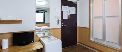 A hotel room with a bed, a desk with a computer, a small refrigerator, a door with a mirror, and a window.