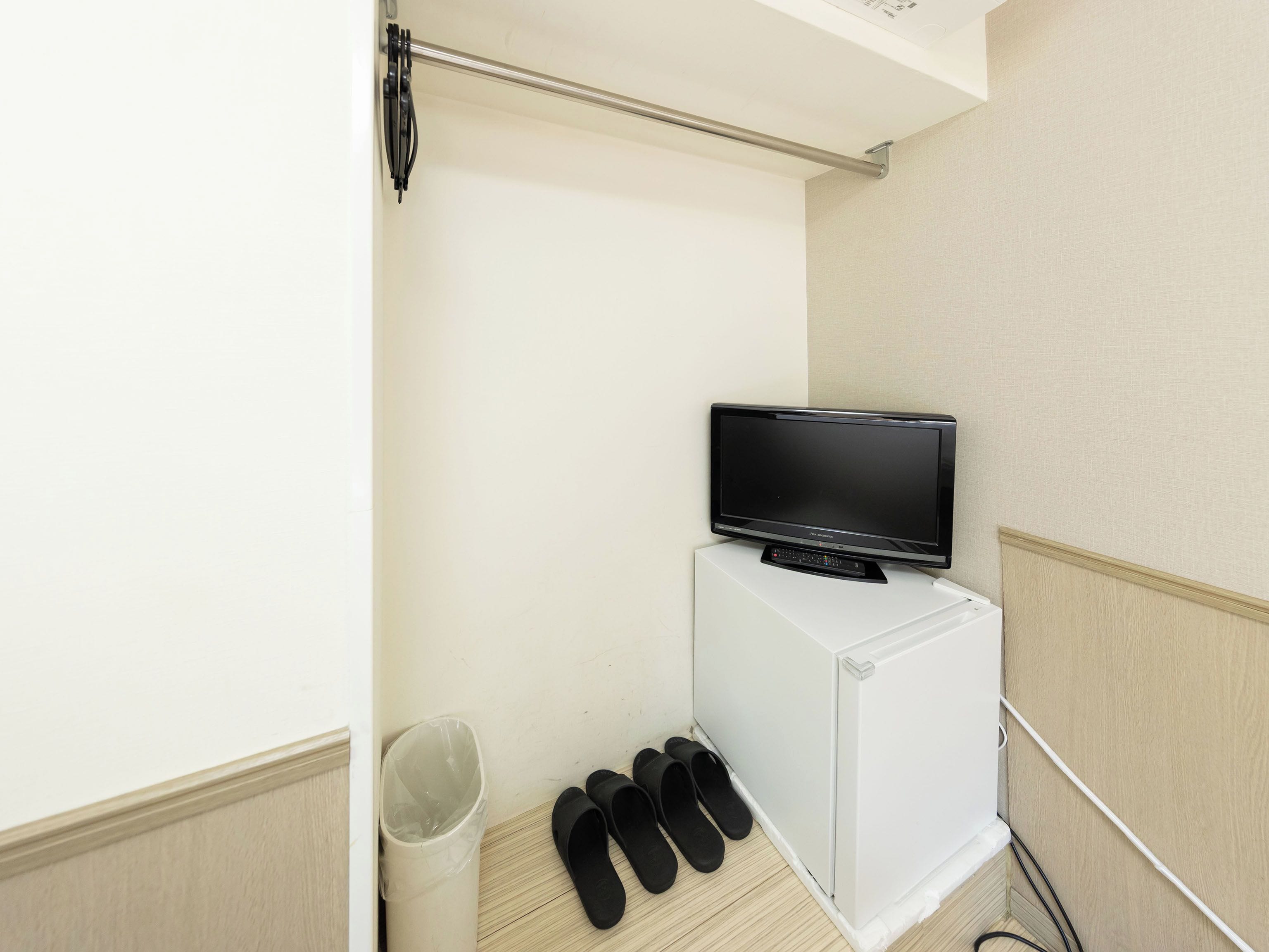 Une petite pièce avec un téléviseur posé sur un meuble blanc, une poubelle et une paire de pantoufles noires.