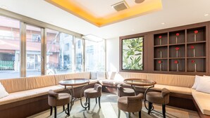 A modern lounge area with a sofa, chairs, a round table, and a large window offering a view of the street. - Tabist Changtee Hotel (Tokyo)