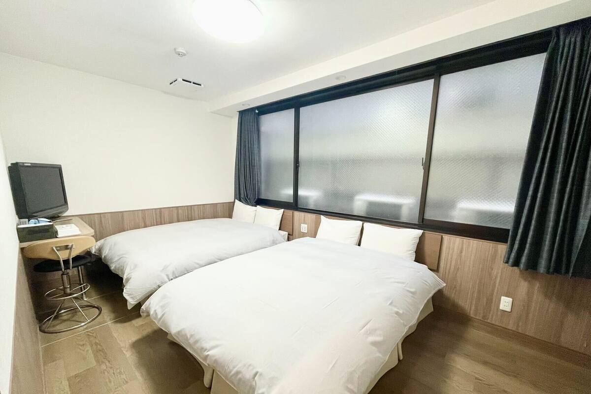 Quarto de hotel com duas camas, uma escrivaninha com televisão e uma janela grande com cortinas.