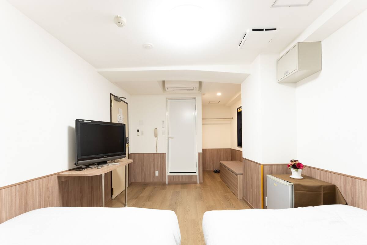 Quarto de hotel com duas camas, televisão, escrivaninha, mini-geladeira, espelho e planta em vaso.