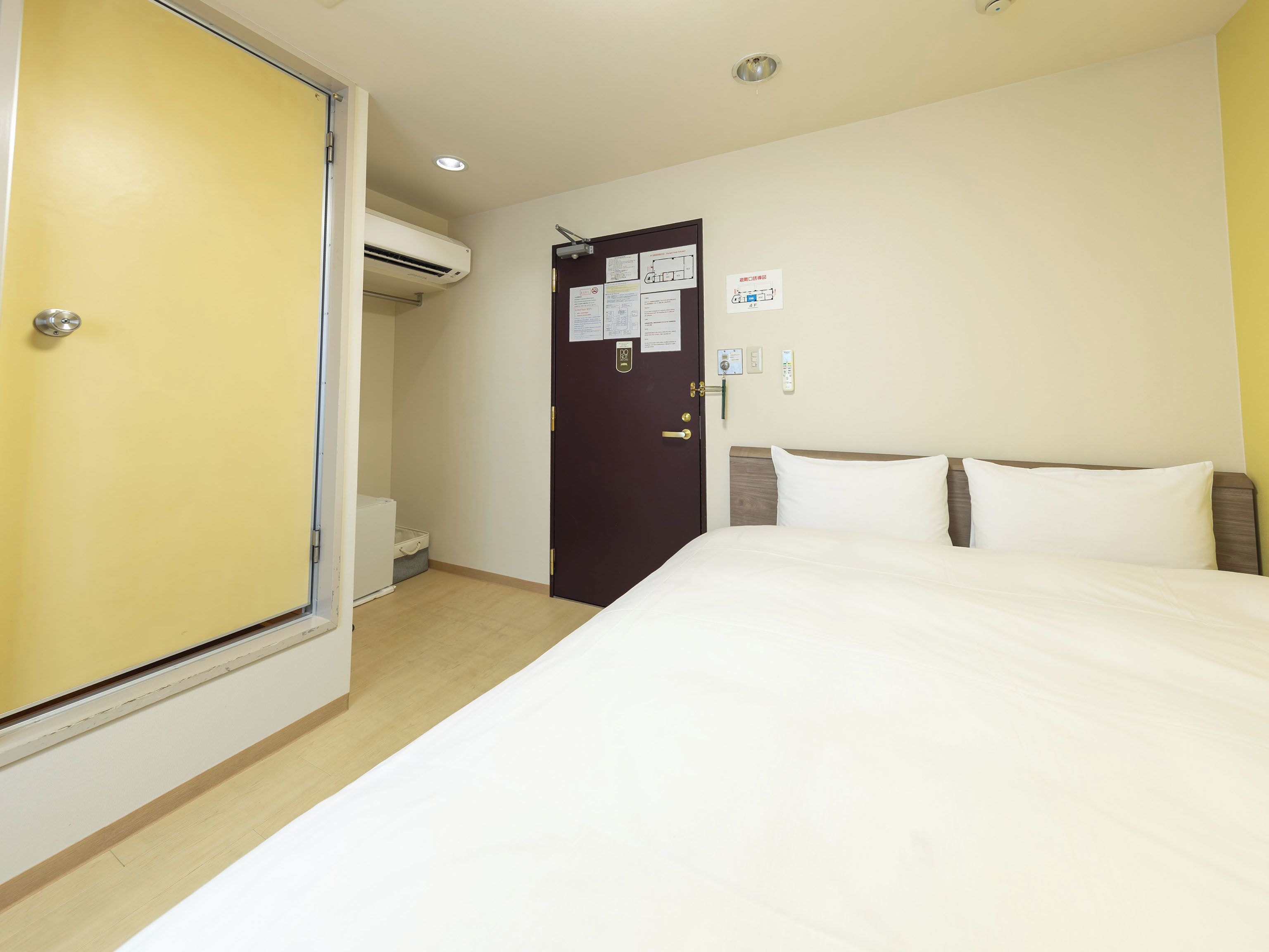 A hotel room with a bed, a door with a lock, an air conditioning unit, and a small desk with a chair.