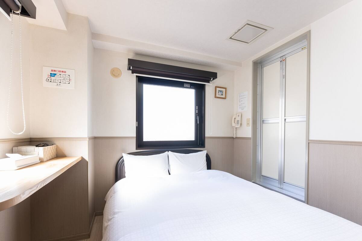 Quarto de hotel com cama, escrivaninha com telefone, janela com persianas e porta com espelho.
