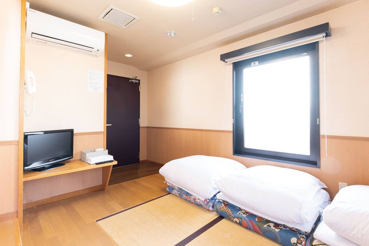 Quarto de hotel com cama, televisão sobre suporte de madeira, escrivaninha com telefone e janela com persianas.