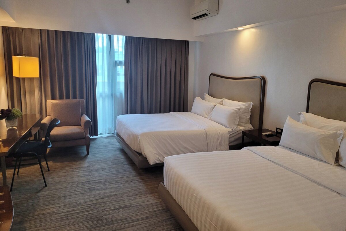 A hotel room with two beds, a desk, a chair, and a lamp.