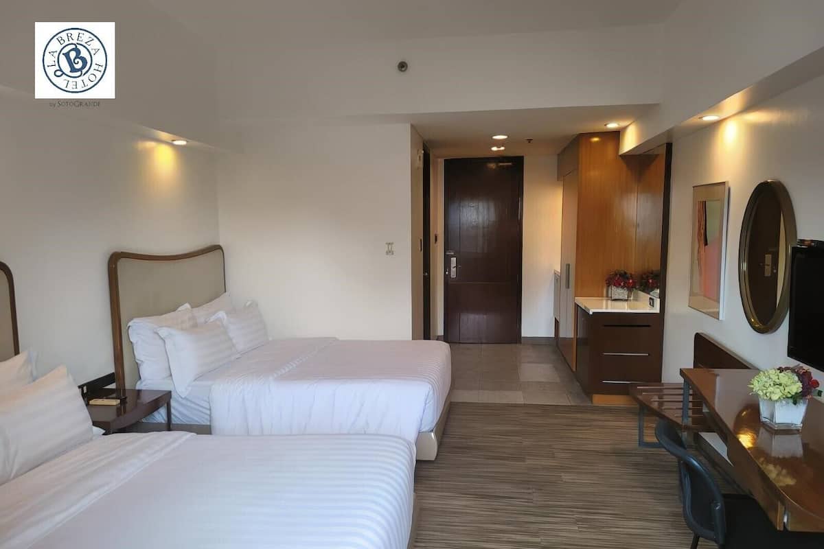 A hotel room with two beds, a desk, a chair, a TV, and a door leading to another room.