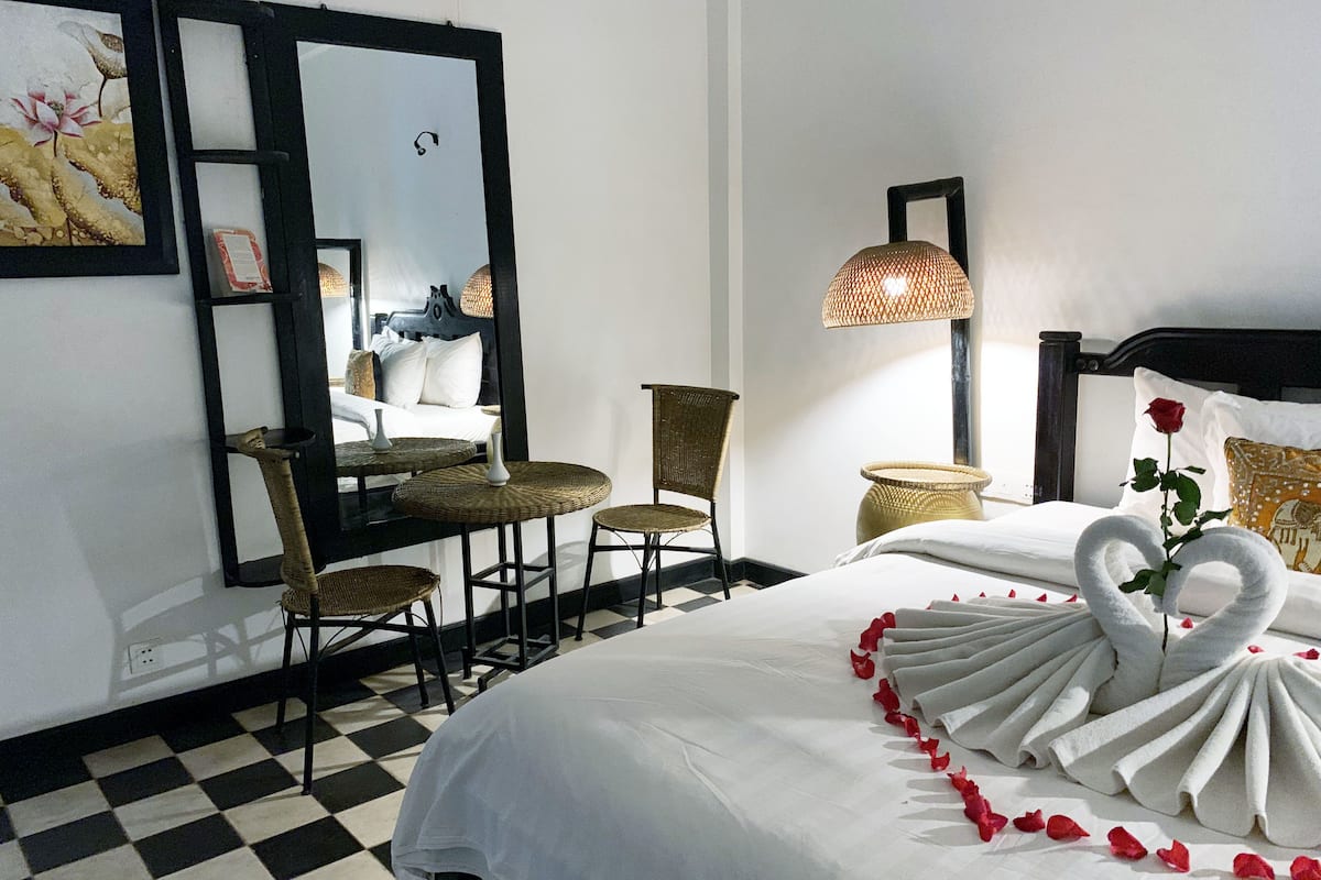 Une chambre d’hôtel avec un sol à carreaux, un lit agrémenté d’une décoration en forme de cygne faite avec des serviettes, un fauteuil en osier, une petite table et un miroir.