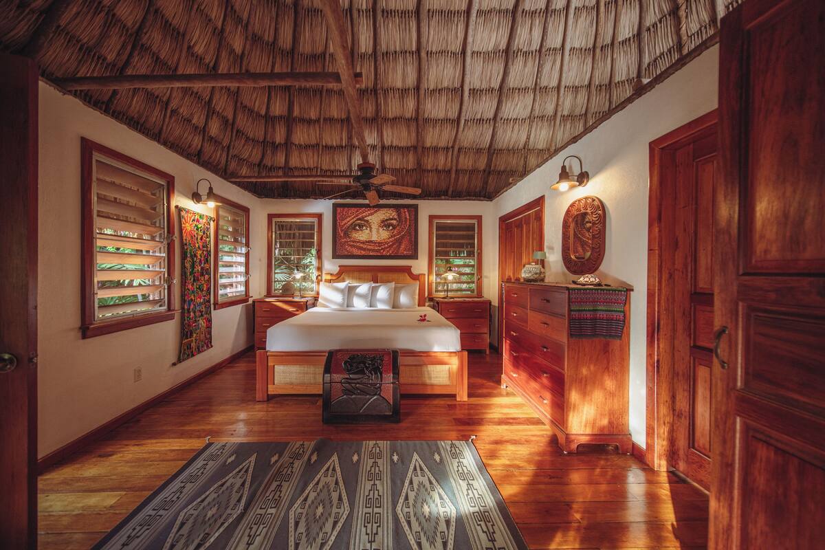 A room with a thatched ceiling, wooden furniture, a bed with white linens, and artwork on the walls.