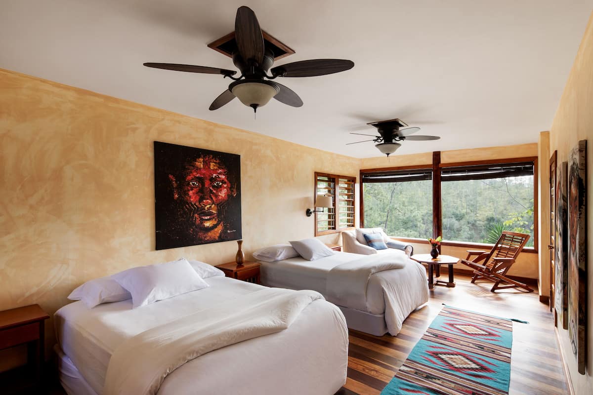 A hotel room with two beds, a ceiling fan, a large window with a view of trees, and a painting on the wall.