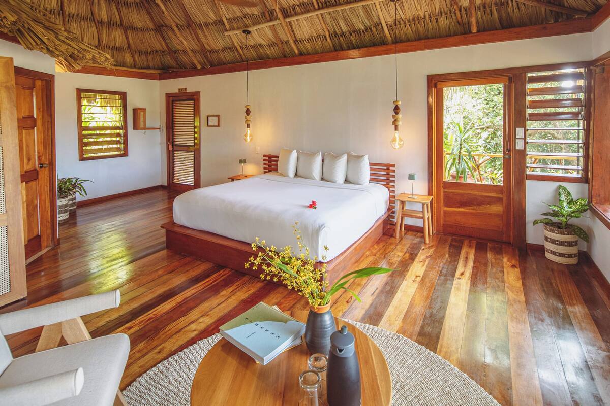 A spacious bedroom with a large bed, wooden flooring, a thatched roof, and a round wooden table with a vase and candles.