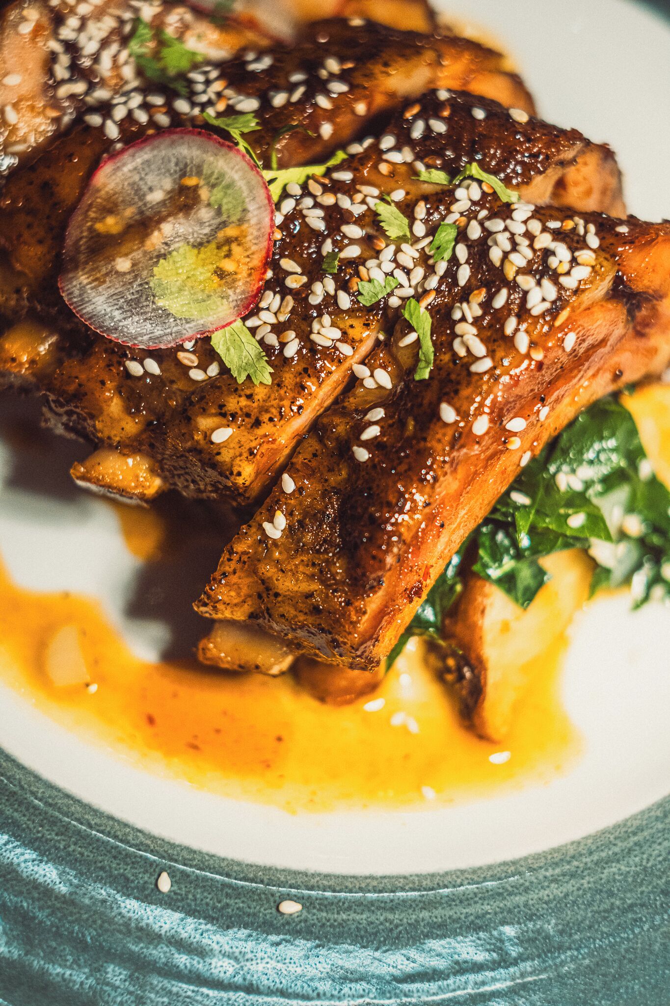 A plate of glazed pork ribs garnished with sesame seeds and herbs, served with a side of greens and a yellow sauce.