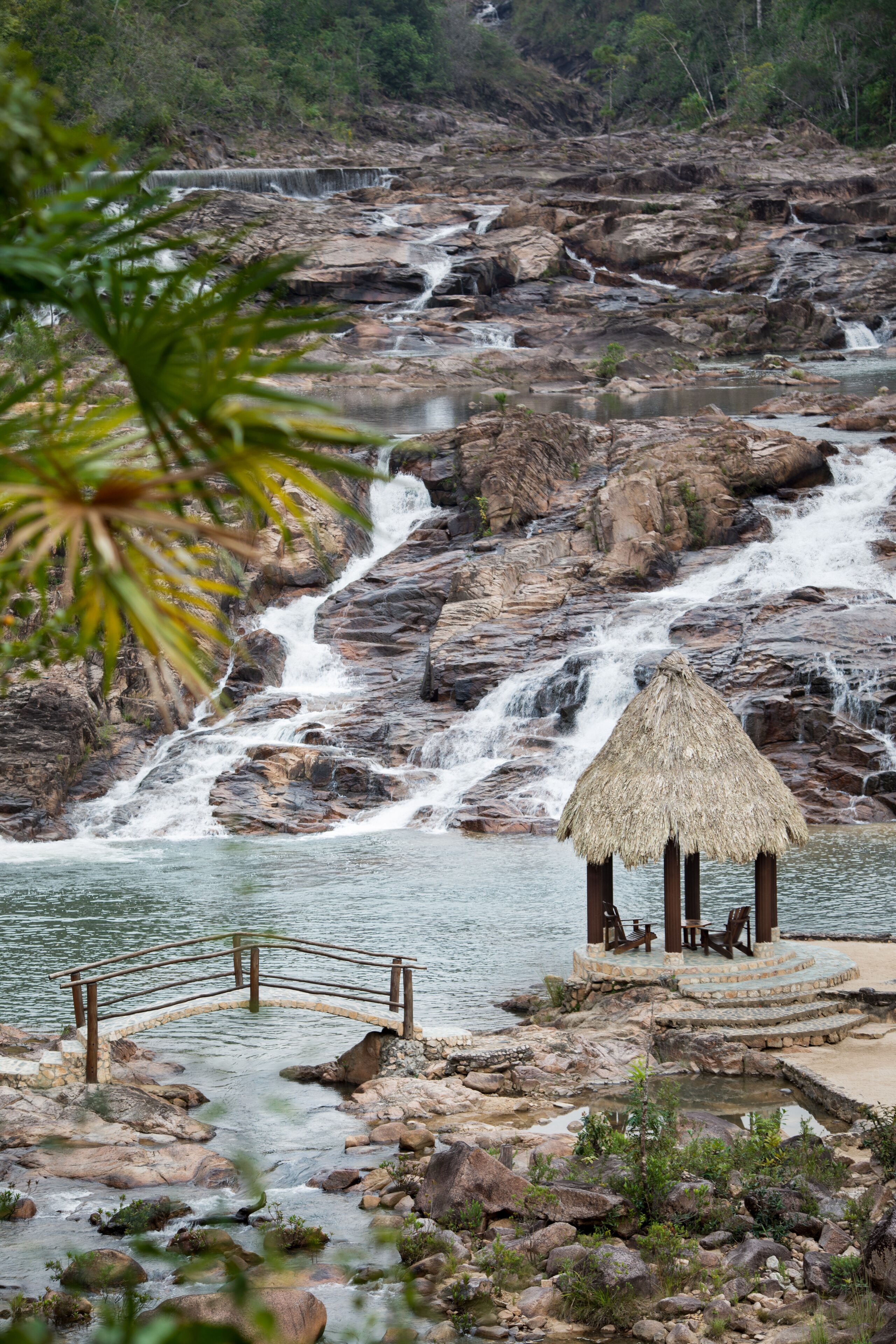 Un paysage rocheux avec une petite construction à toit de chaume et un pont en bois enjambant une étendue d’eau.