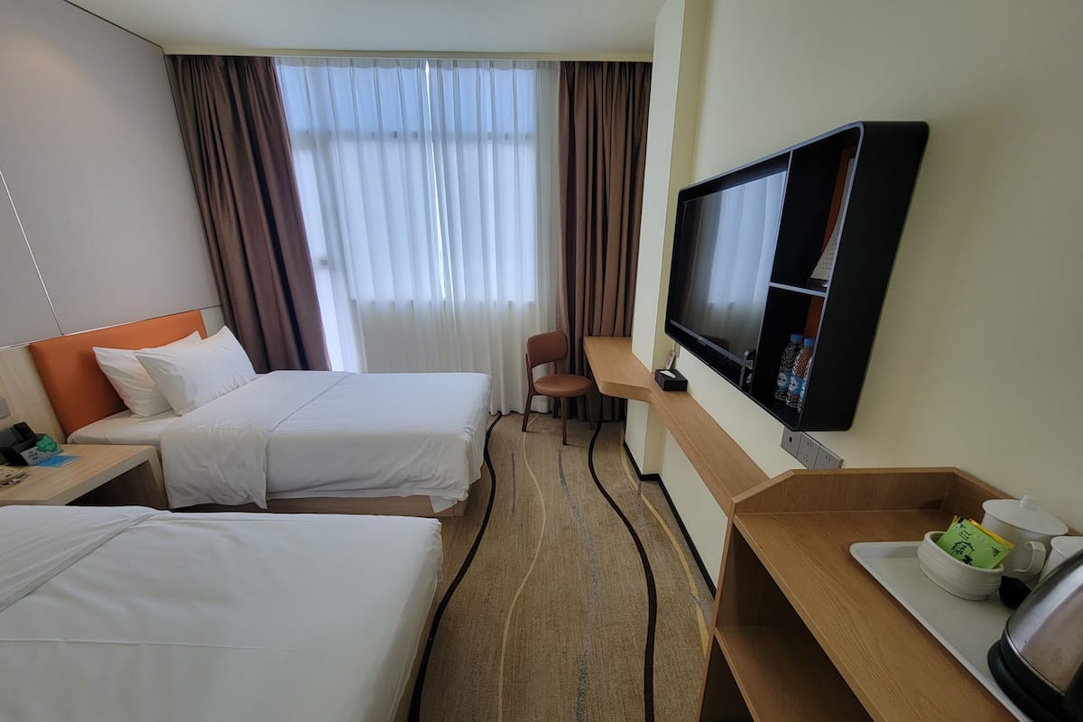 A hotel room with two beds, a desk with a television, a chair, and a tray with a kettle and cups.