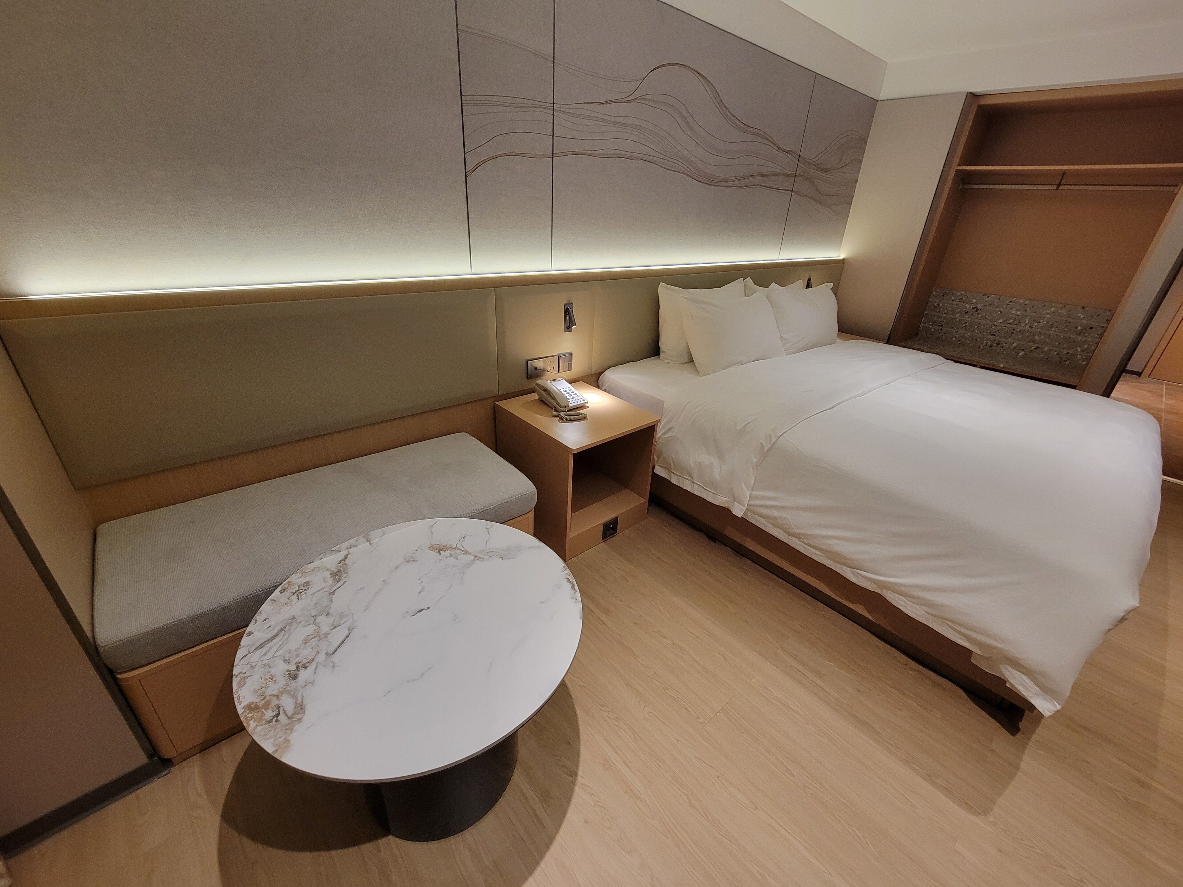 A compact hotel room with a bed, a bedside table with a phone, a small round table, and a bench.