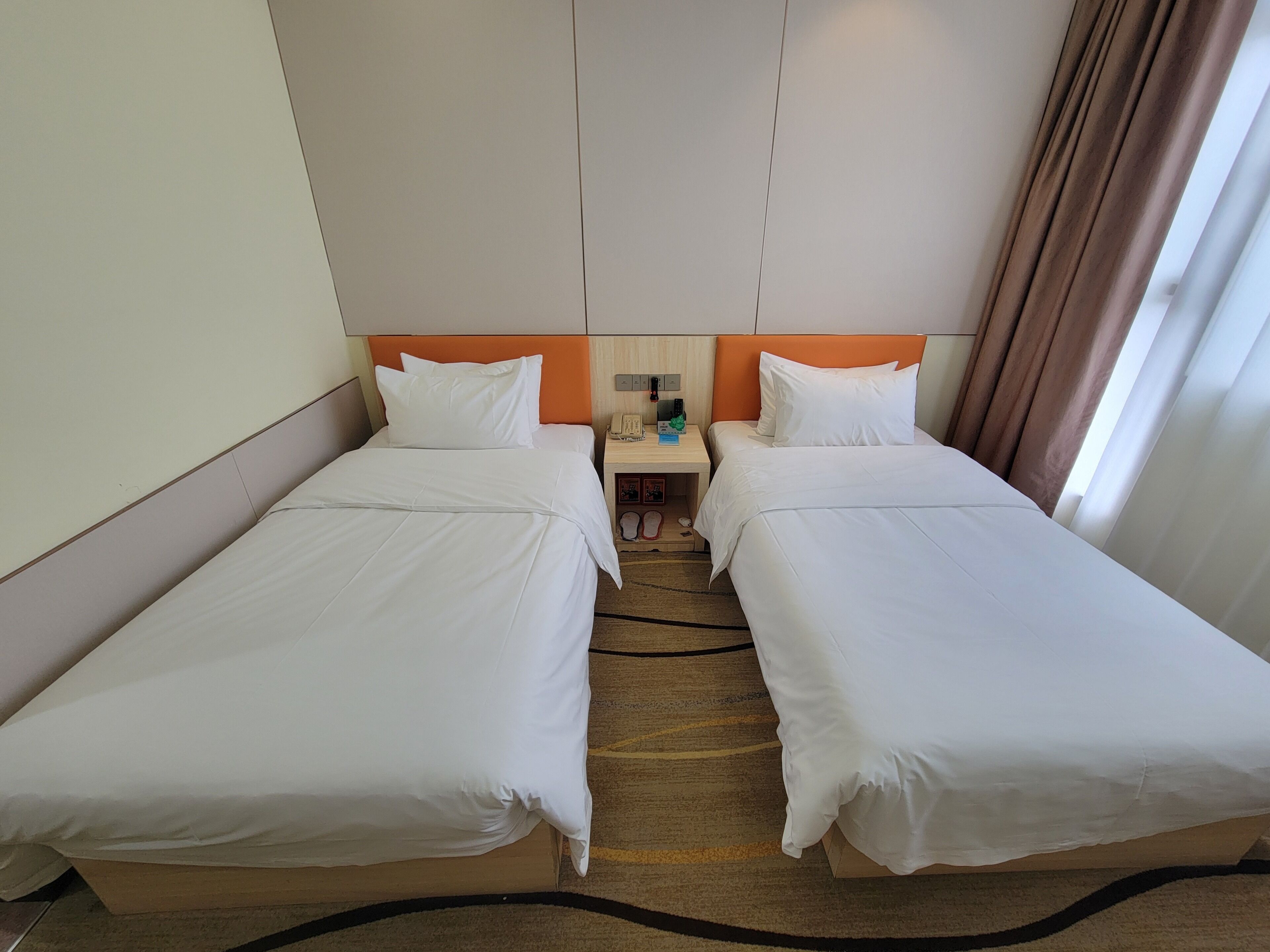 Habitación de hotel con dos camas individuales, una mesita de noche con teléfono y lámpara, y vista a la calle por la ventana.