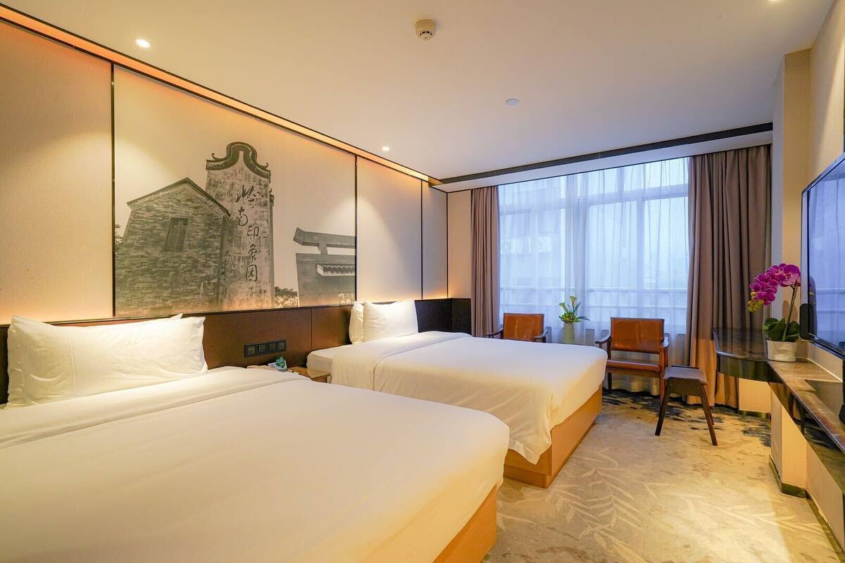 Une chambre d’hôtel avec deux lits, un bureau avec une chaise, une grande fenêtre avec des rideaux et un élément décoratif mural.