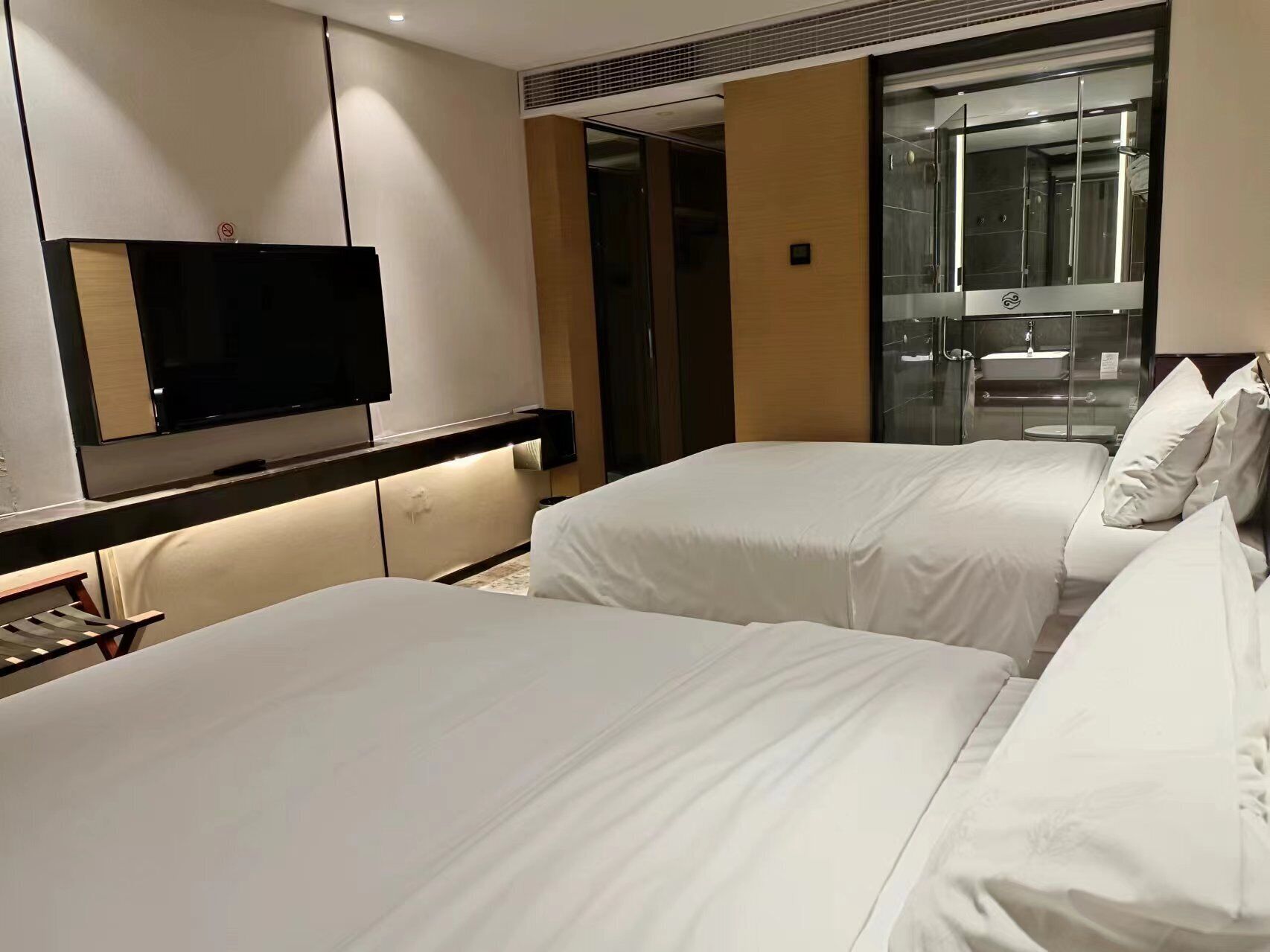 A hotel room with two beds, a TV, a glass-enclosed bathroom, and a wooden panel wall.