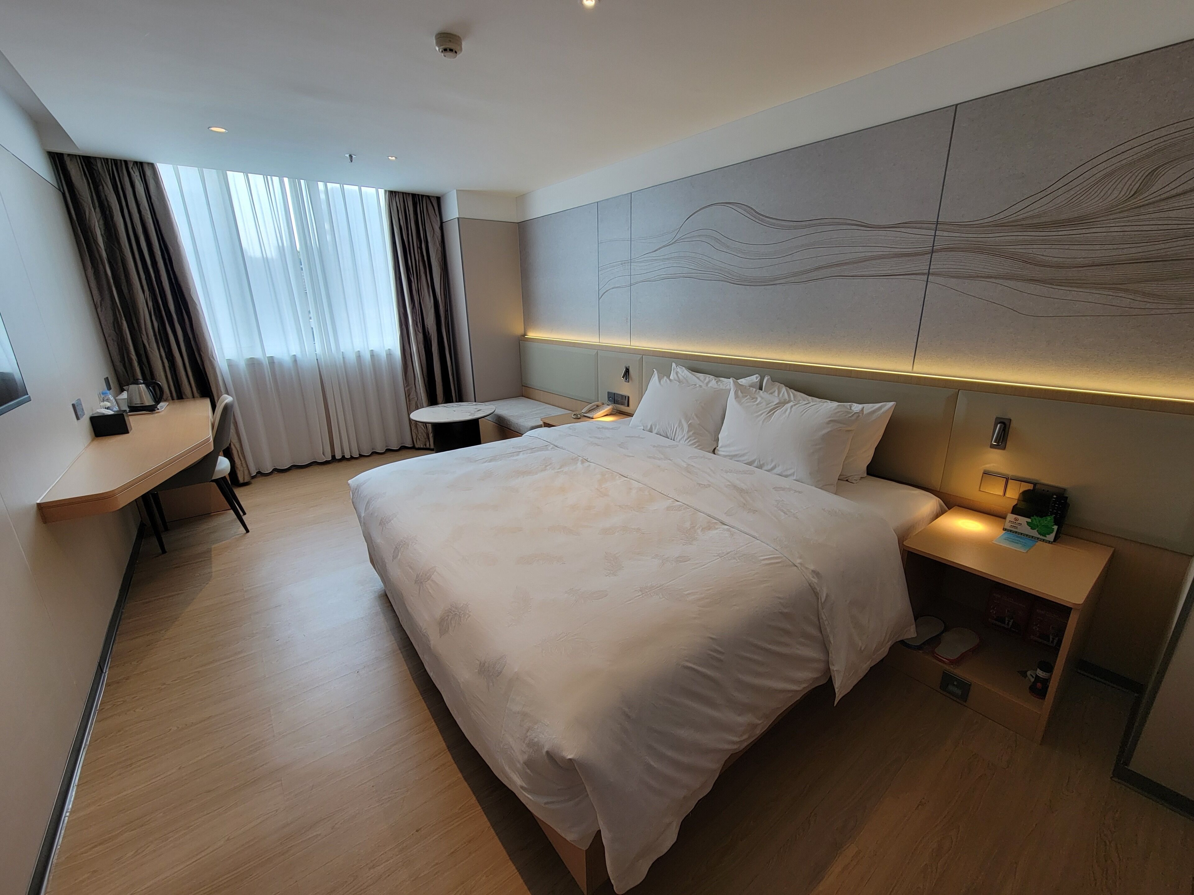 A modern hotel room with a large bed, a desk, and a view of the outdoors through the window.