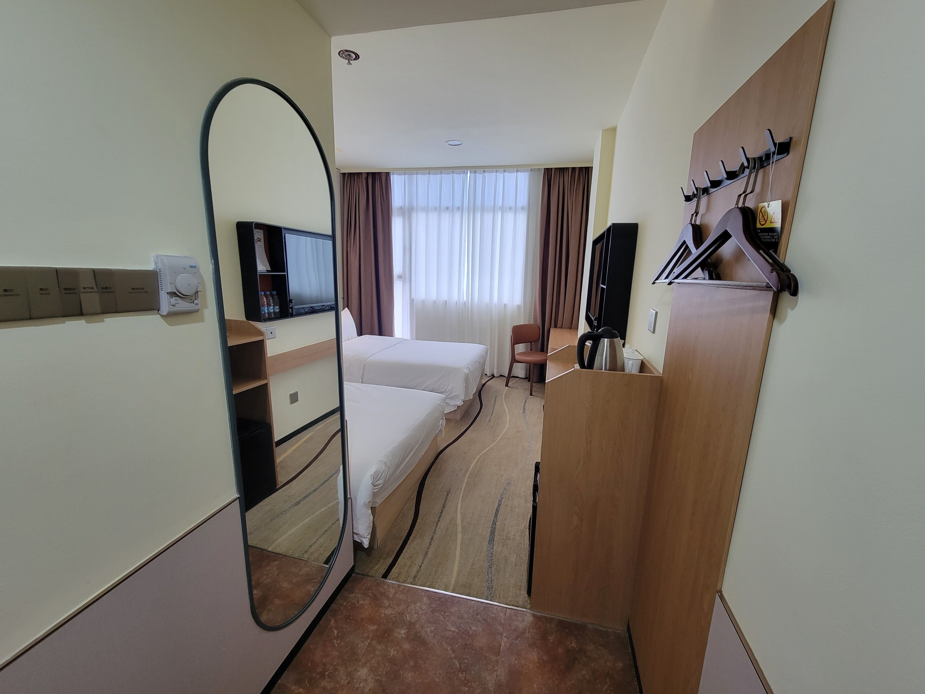 A hotel room with a bed, a desk with a chair, a television, a mirror, and a coat rack.