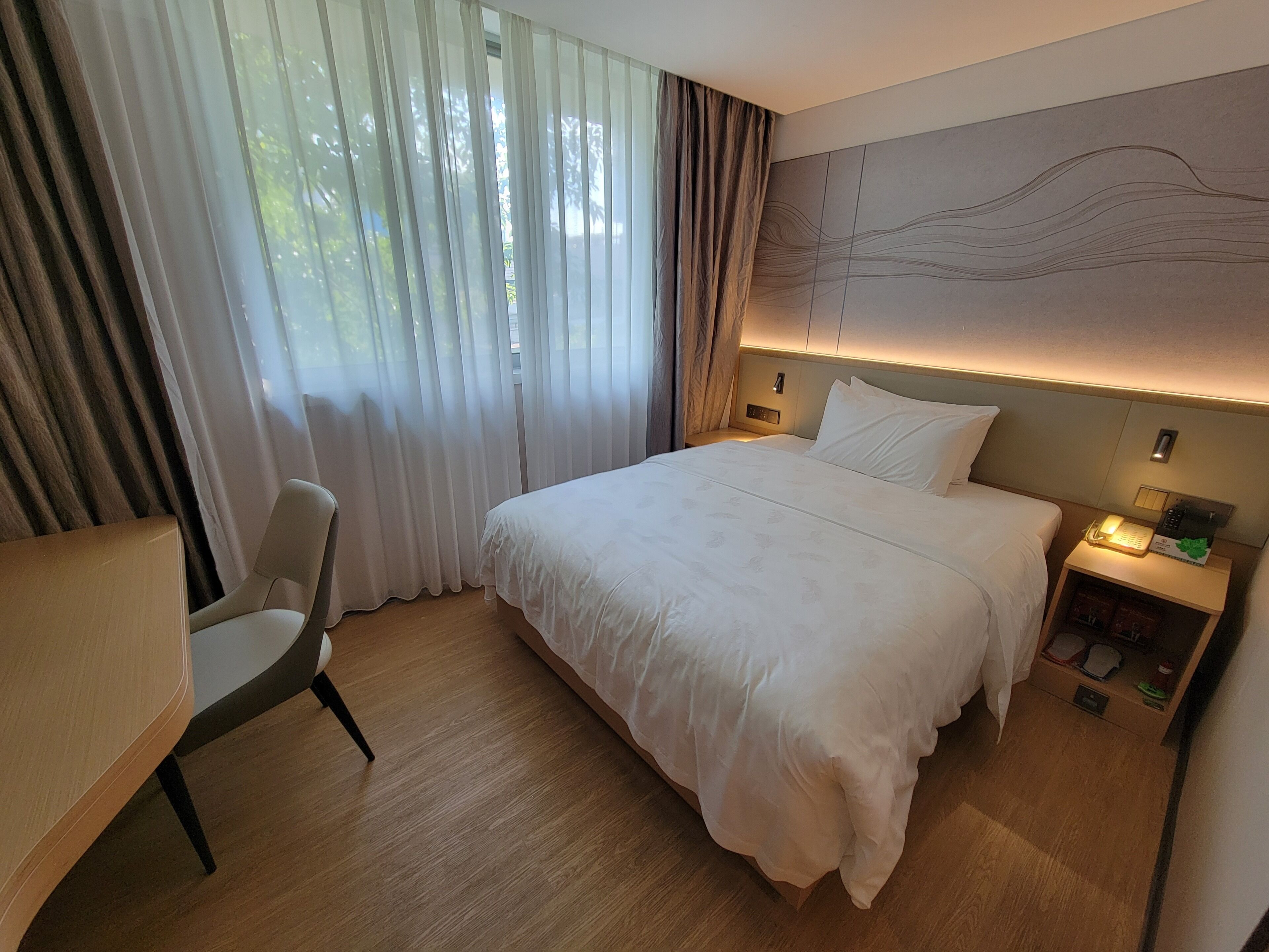 A hotel room with a large bed, a desk, a chair, a nightstand with a phone and a lamp, and a view of trees through sheer curtains.