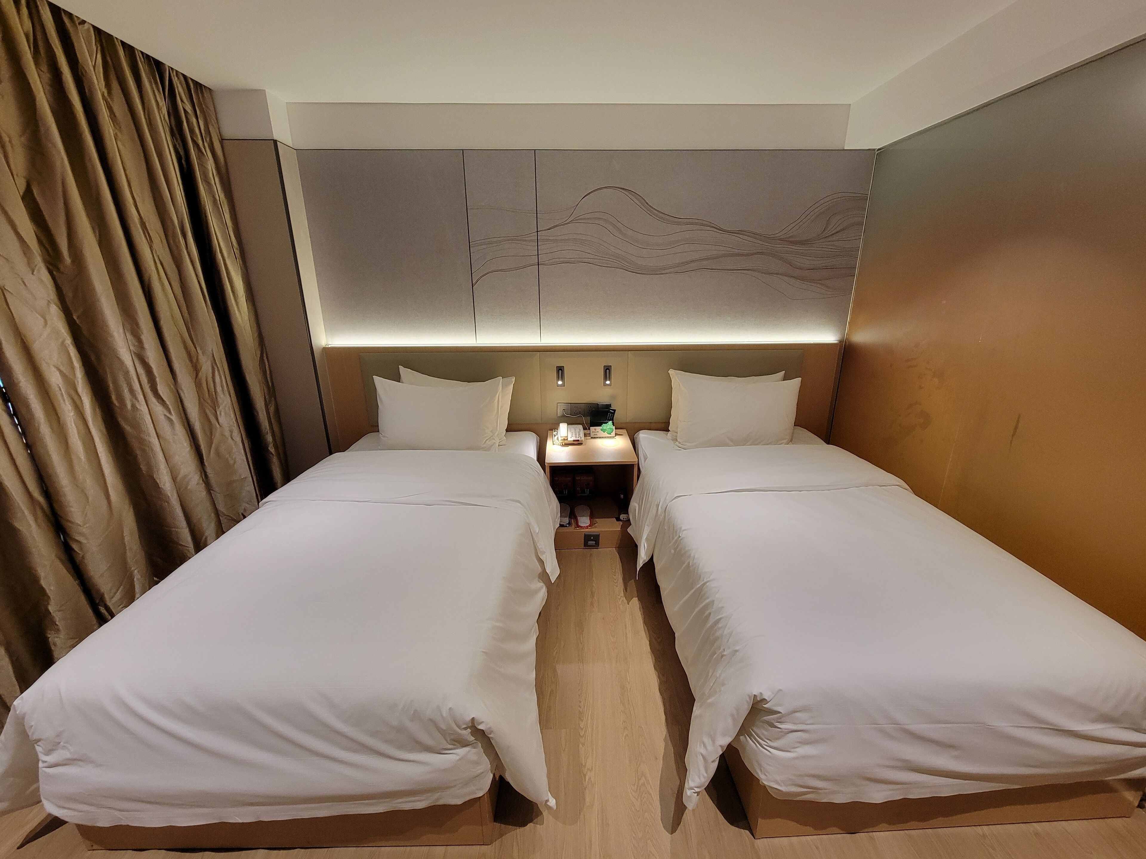 A hotel room with two single beds, a bedside table with a lamp, and a wall-mounted artwork.
