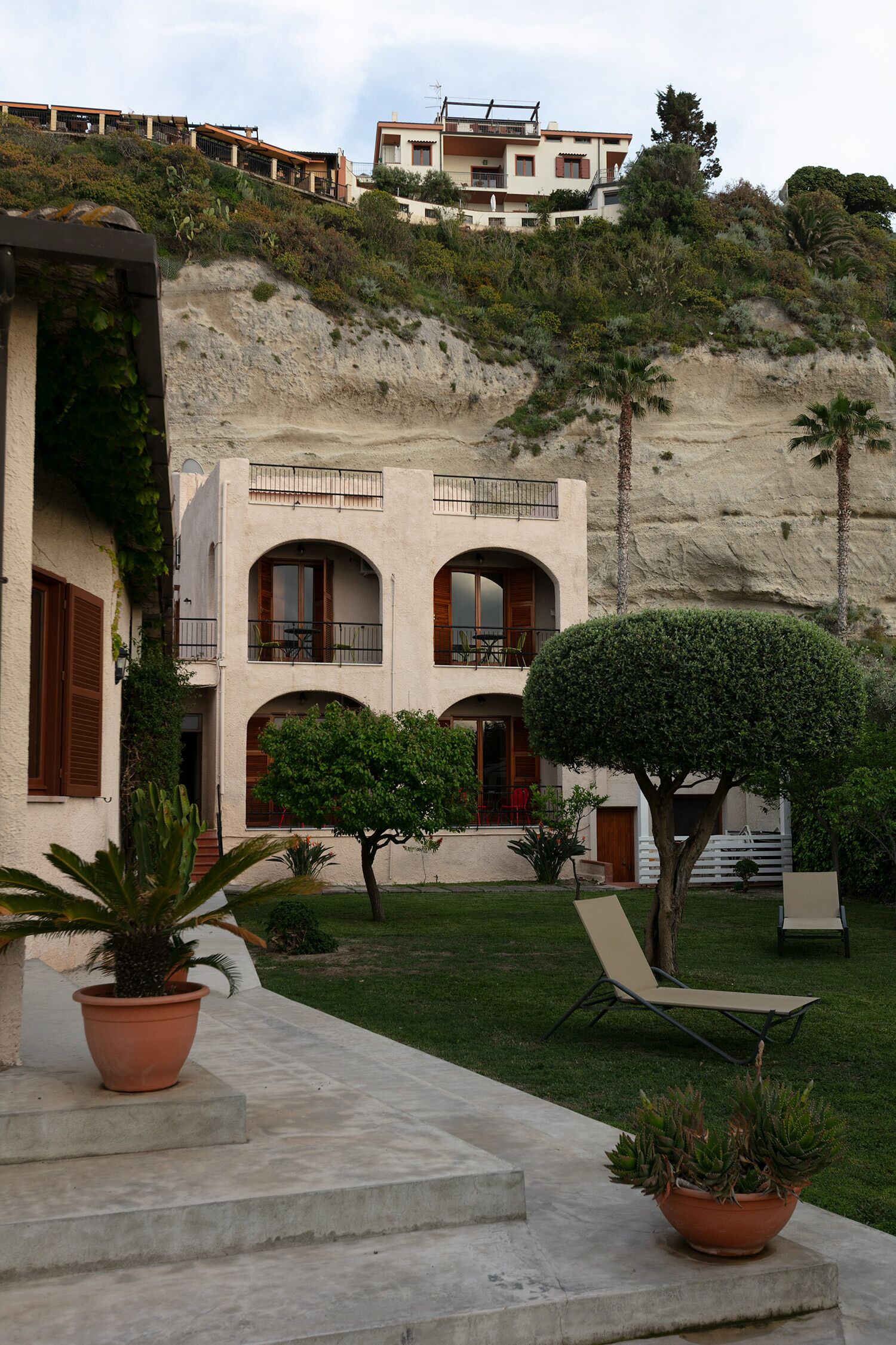 A two-story building with a courtyard, featuring a staircase leading to the entrance, a small pool area, and a view of a hillside.