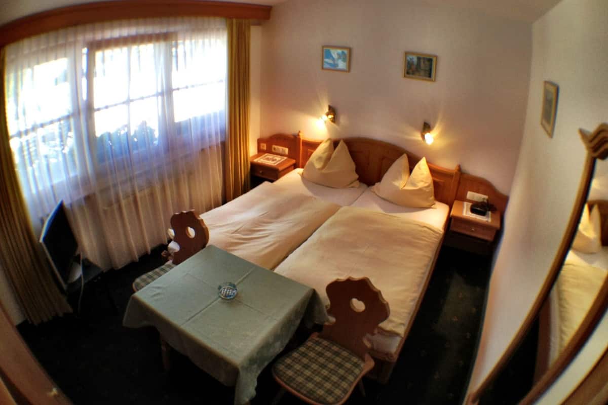 A hotel room with two beds, a table, and a chair. There are two bedside tables with lamps, a window with curtains, and two small wall art pieces.