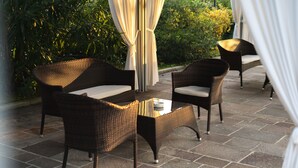 An outdoor patio with wicker furniture and a glass table, surrounded by greenery and stone flooring.