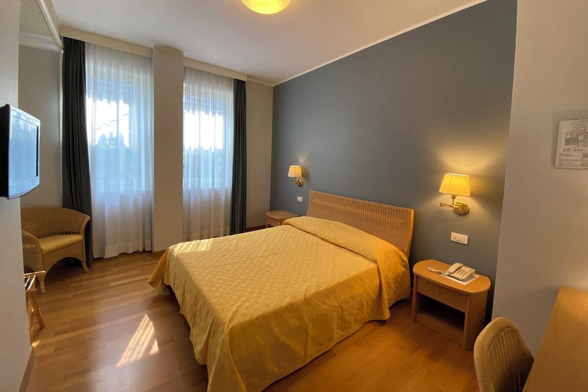 A hotel room with a bed, two bedside lamps, a small table with a phone, a chair, and a television on the wall.
