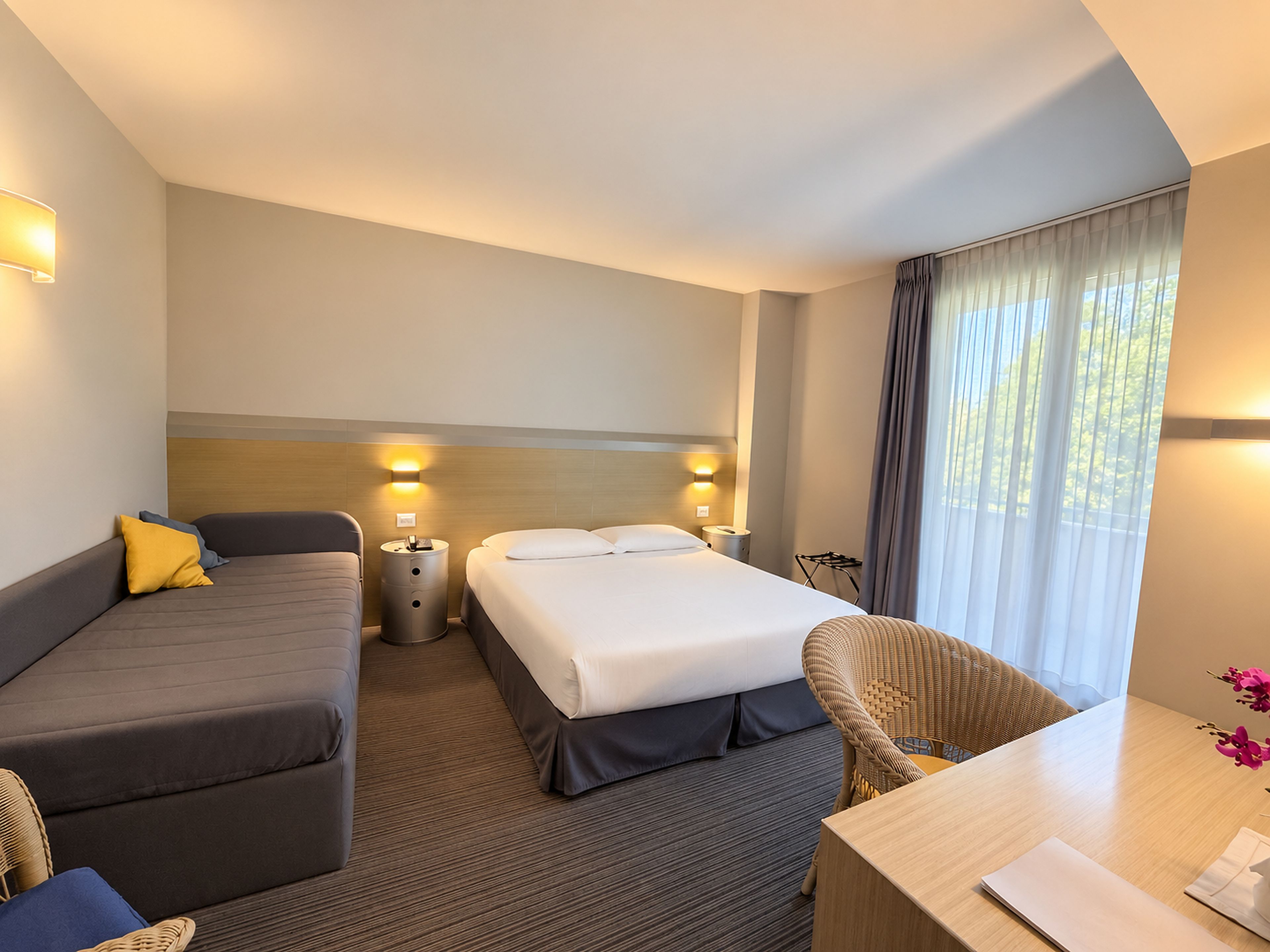 Habitación de hotel con cama, sofá, silla, una mesita y ventana con cortinas.