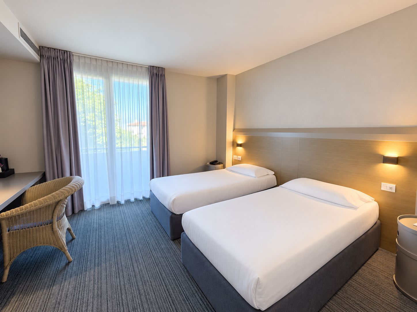 A hotel room with two beds, a desk, a chair, and a view of the outdoors.