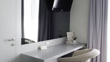 Et moderne hotelværelse med et stort spejl, et fladskærms-tv, et hvidt skrivebord, en fletstol og et stort vindue med gardiner.