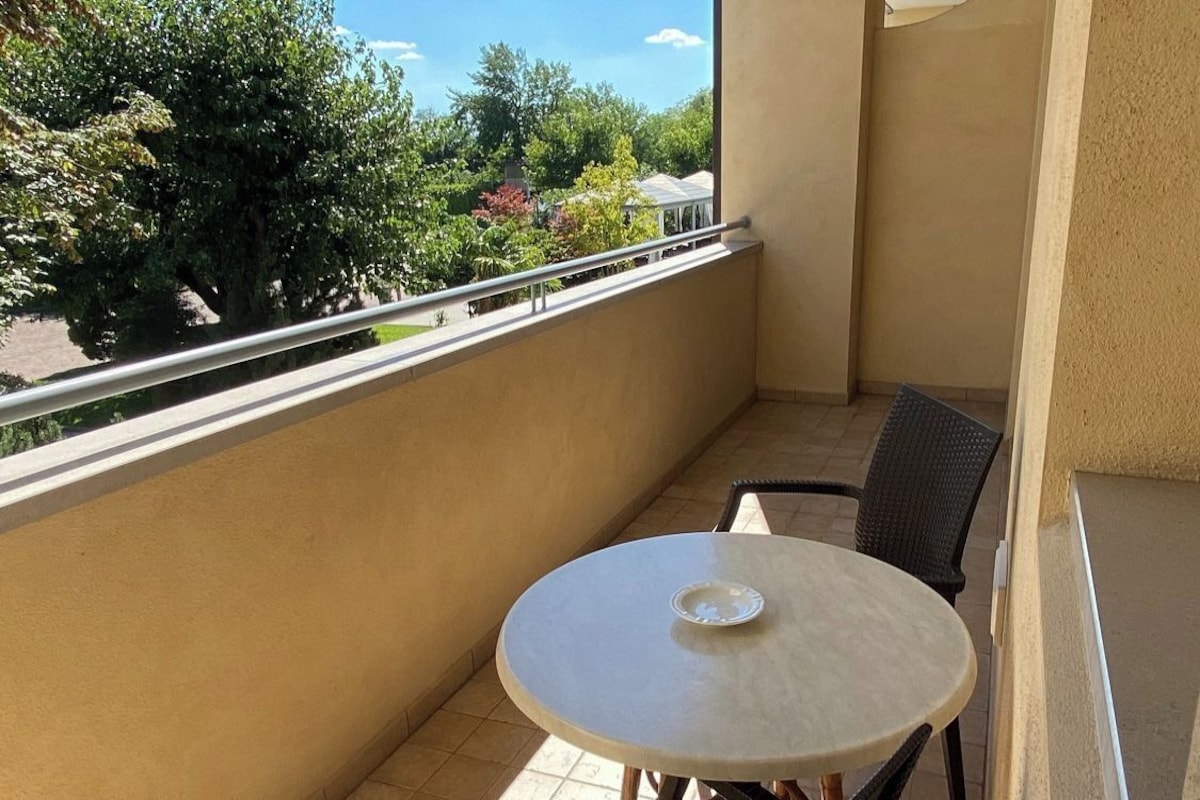 Un balcon avec une table ronde et une chaise, une balustrade et une vue sur les arbres et les bâtiments.