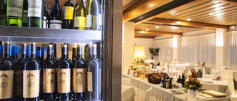 Un refrigerador de vinos repleto de diversas botellas, un comedor con mesas dispuestas para invitados, y un techo con vigas de madera.